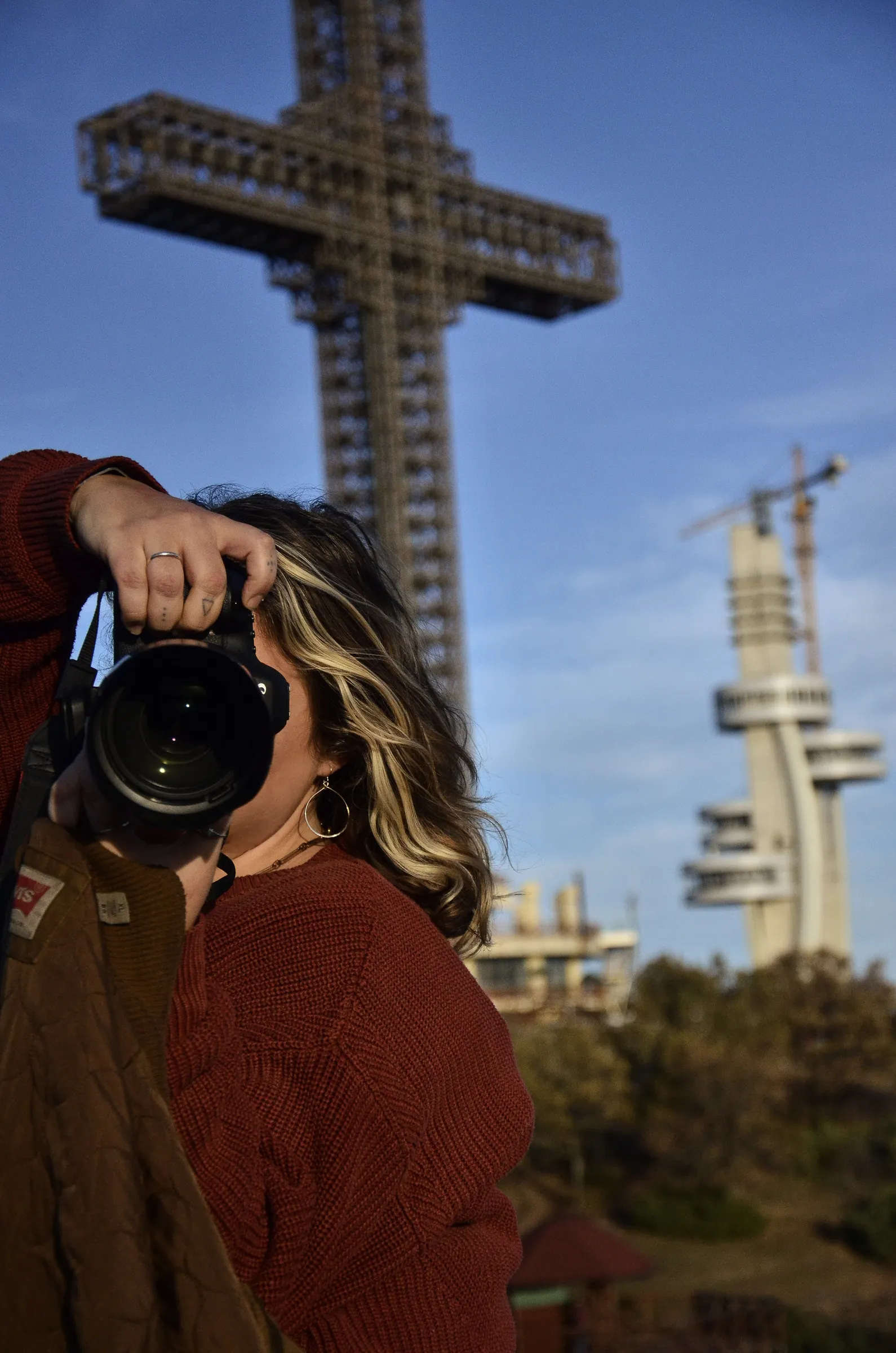 Alex aiming her camera straight at the viewer, the Millennium Cross on Vodno above Skopje behind her