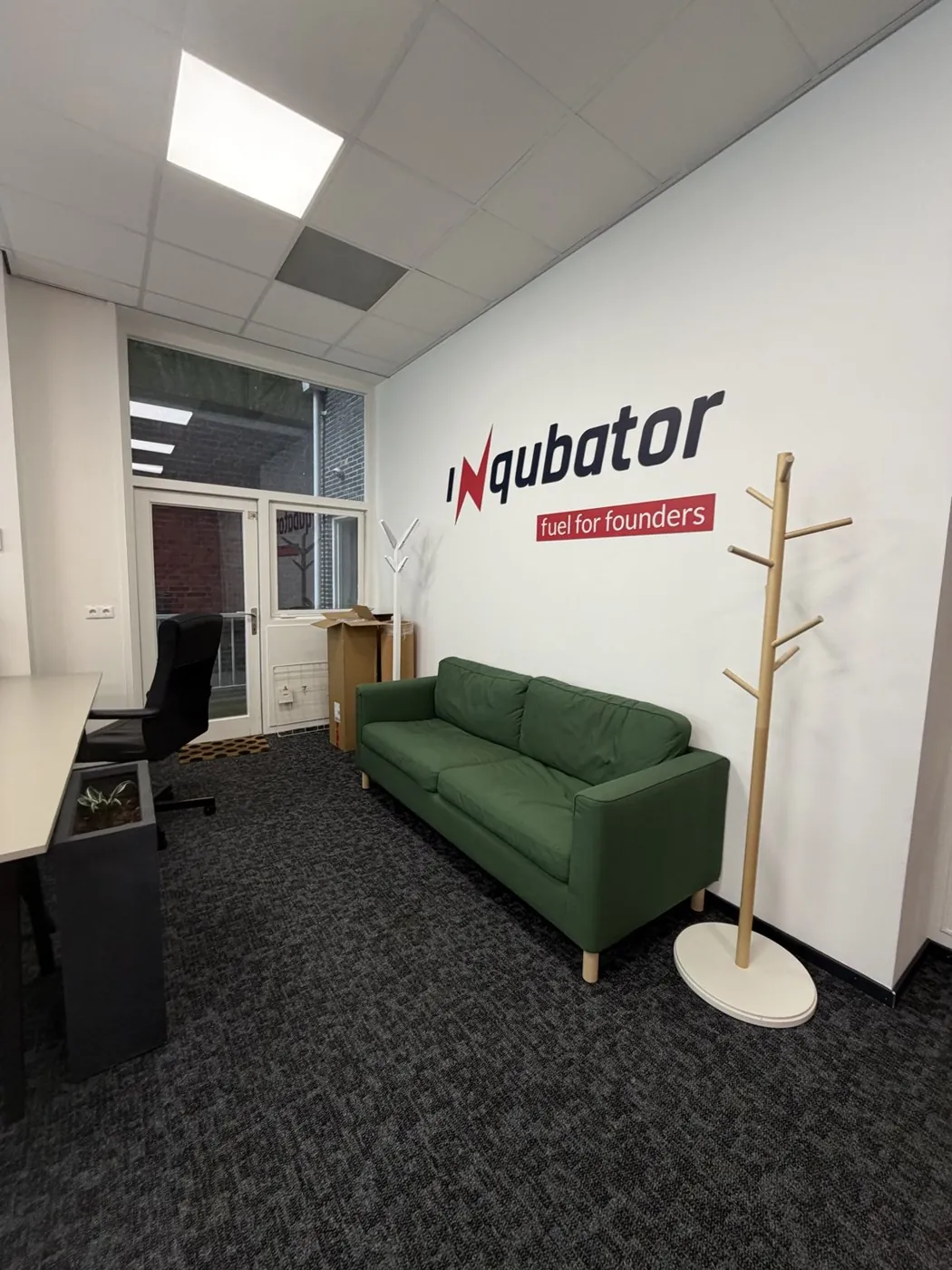 The front of the room in February 2026: a green sofa, a coat stand and the Inqubator logo on a white wall