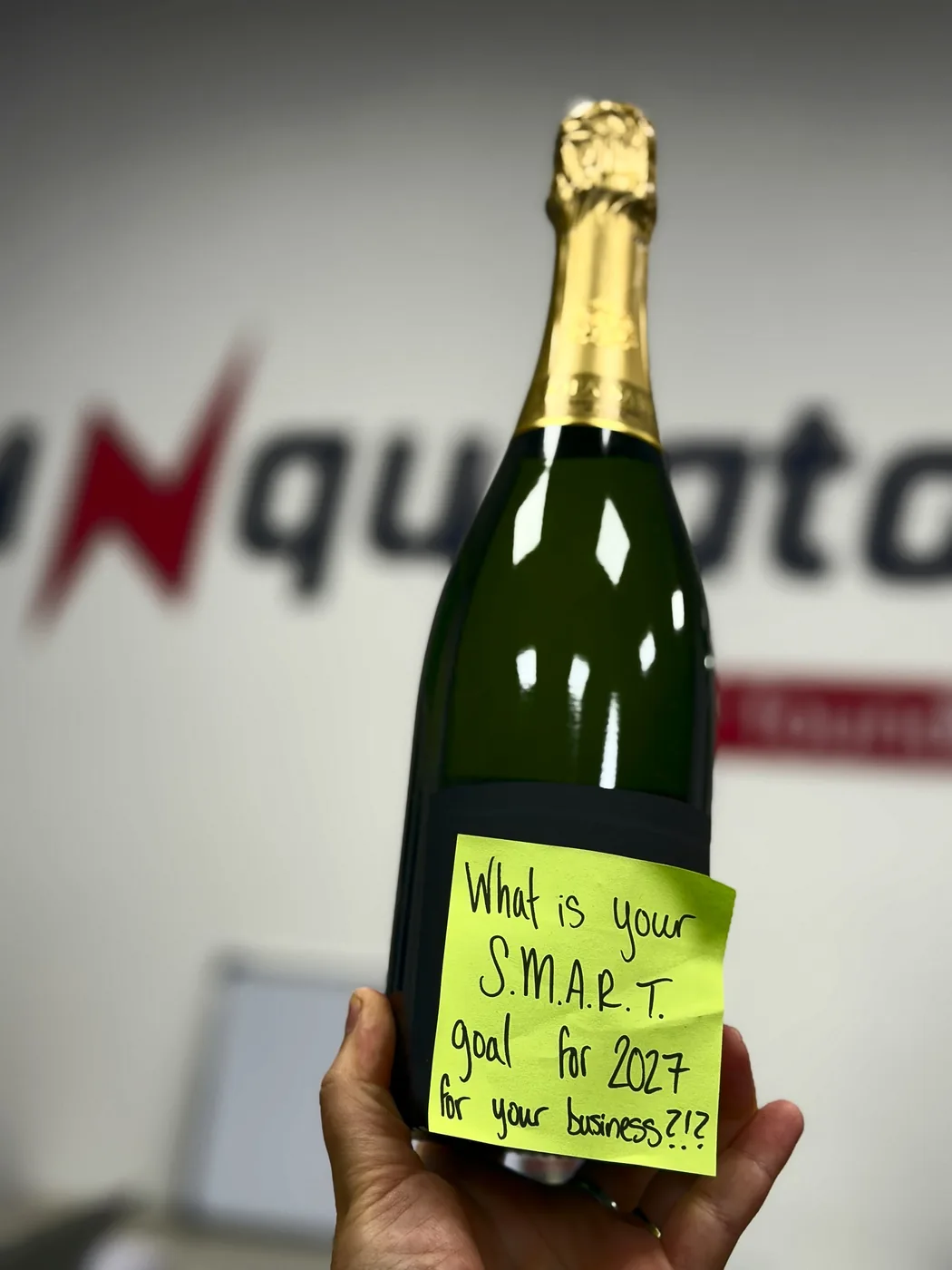 A hand holding a champagne bottle with a sticky note: what is your SMART goal for 2027 for your business?