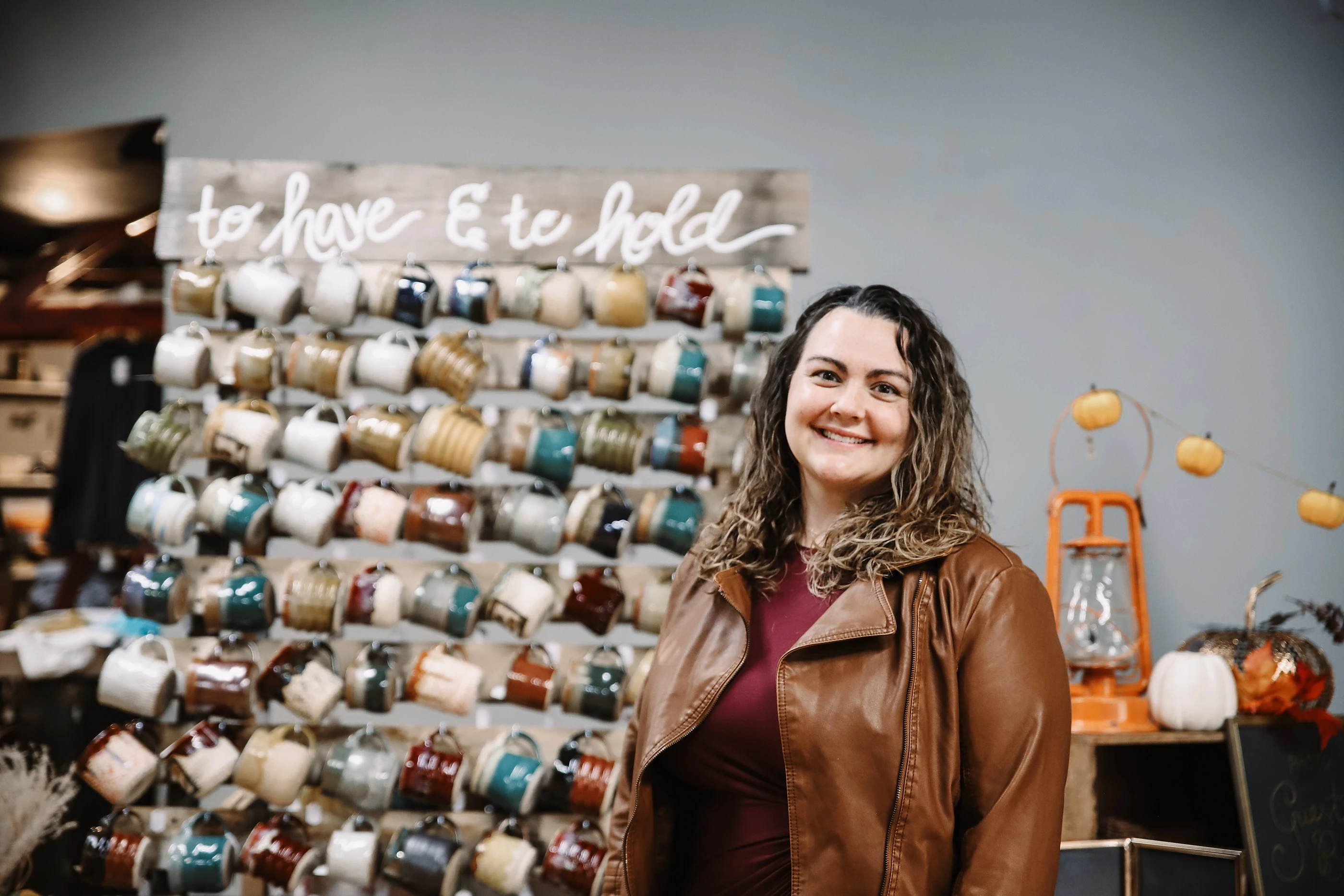 Abby smiling in front of her wall of handmade mugs, under a sign that says to have and to hold