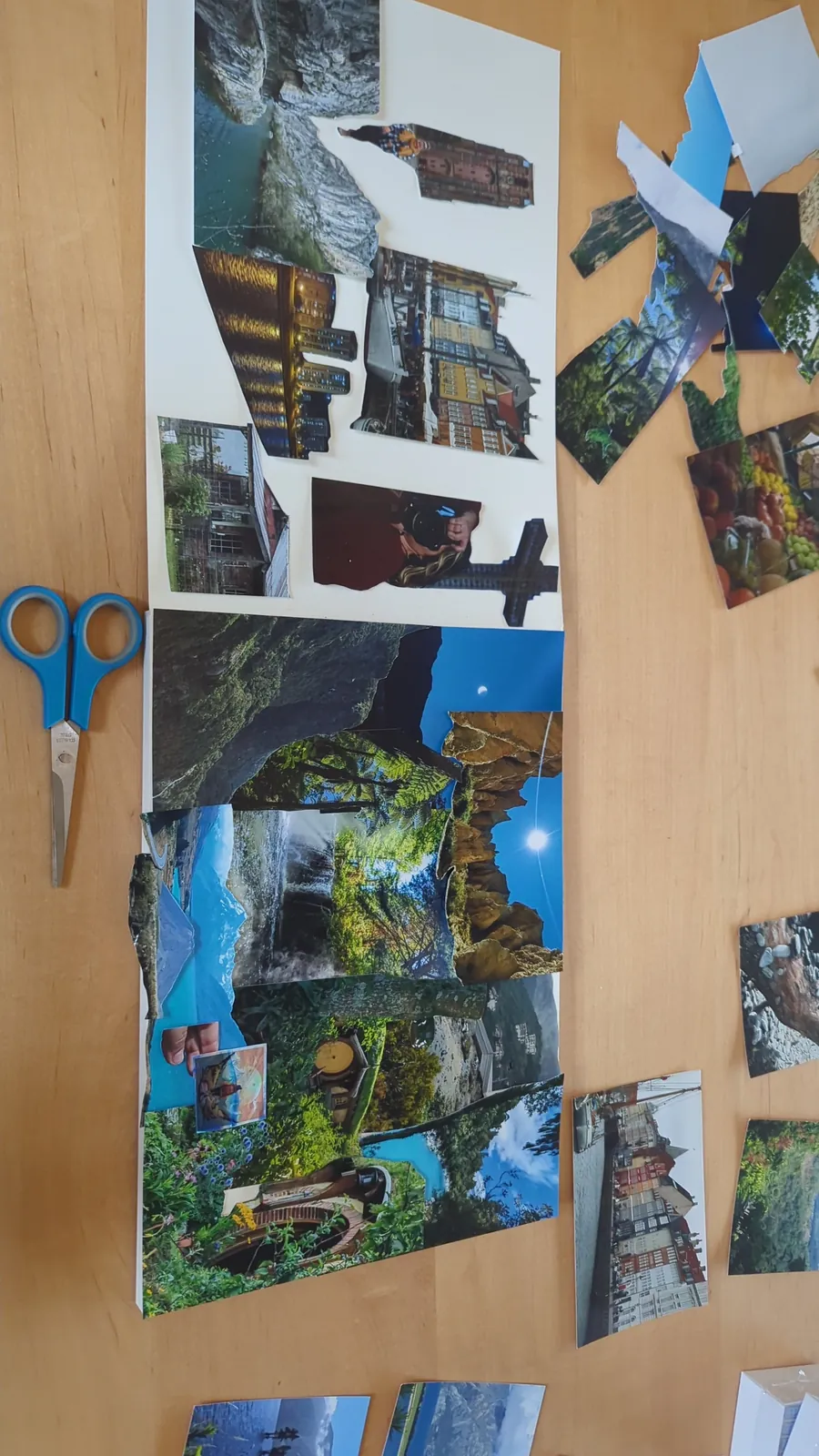 Travel photos cut out by hand and spread across a wooden table, with a pair of blue scissors