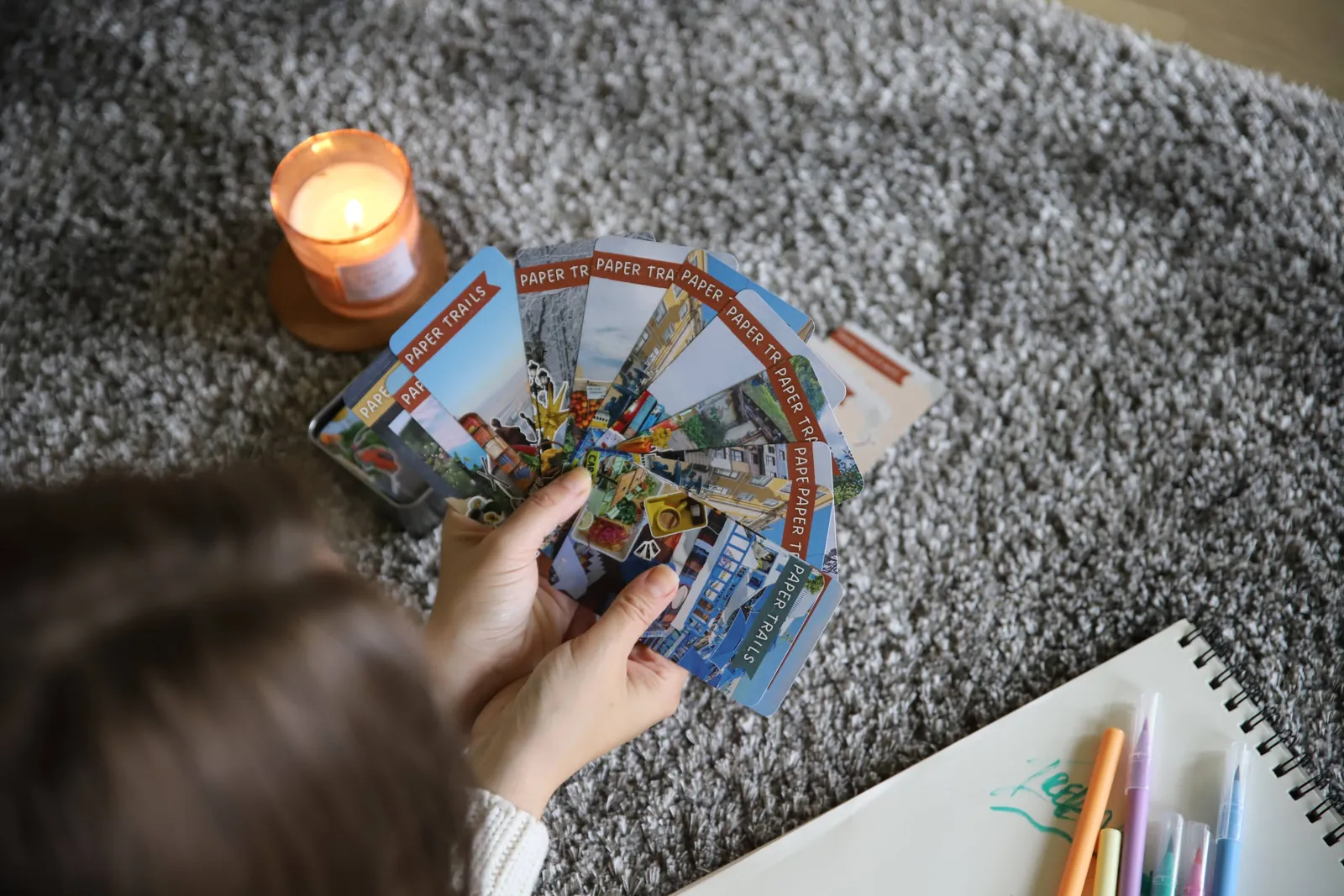 A hand fans out Paper Trails cards over a grey rug, next to a lit candle and a sketchbook