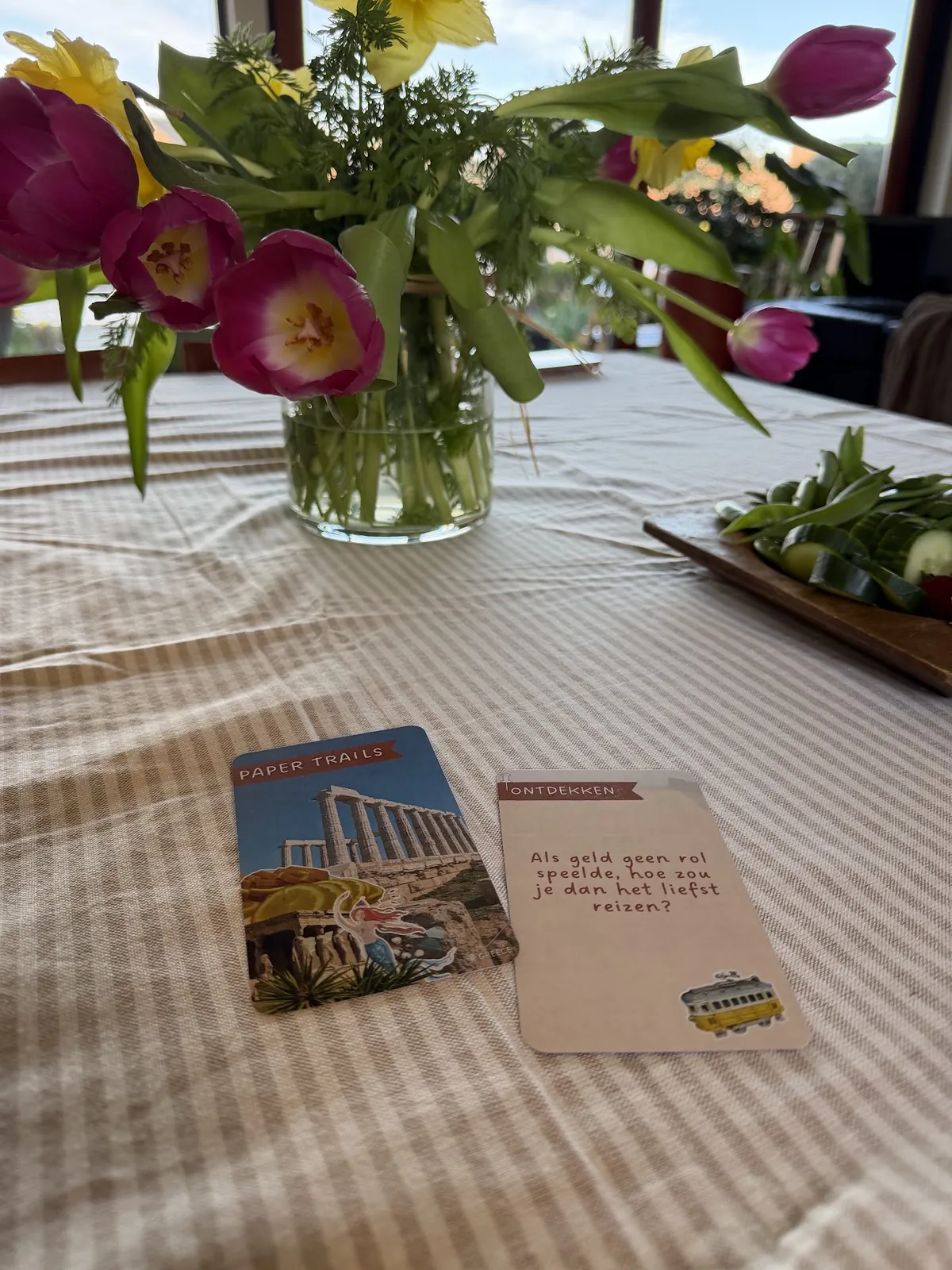 A card front with a Greek temple next to a Dutch question card on a striped tablecloth, with tulips behind