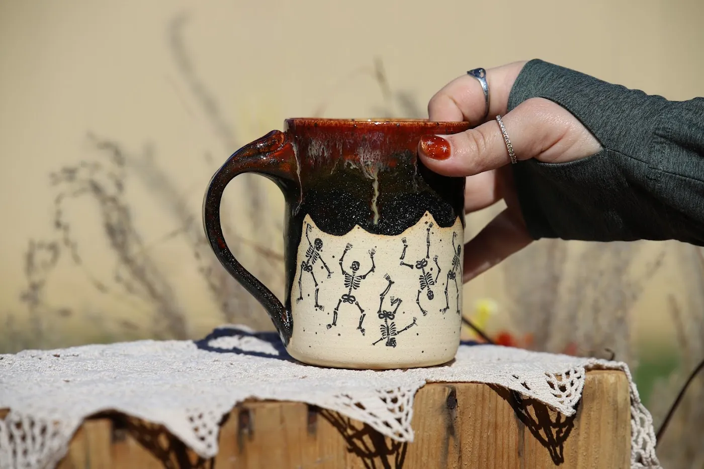 A hand holding a mug painted with dancing skeletons under a dark red glaze